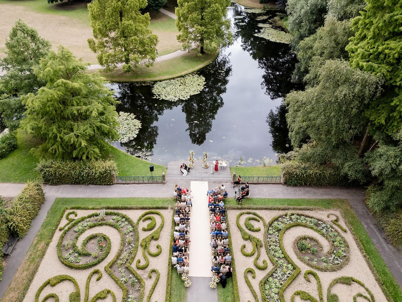 Luchtfoto van een buitenhuwelijk in een formele tuin met sierhagen en een vijver, met gasten in rijen rond het bruidspad.